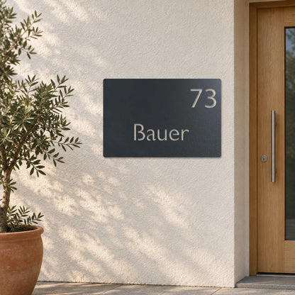 Edelstahl Hausnummernschild personalisiert – Moderne Nummer- und Namensschild „73 Bauer“ an heller Putzwand, Edelstahl Anthrazit RAL 7016 · WesterWerk